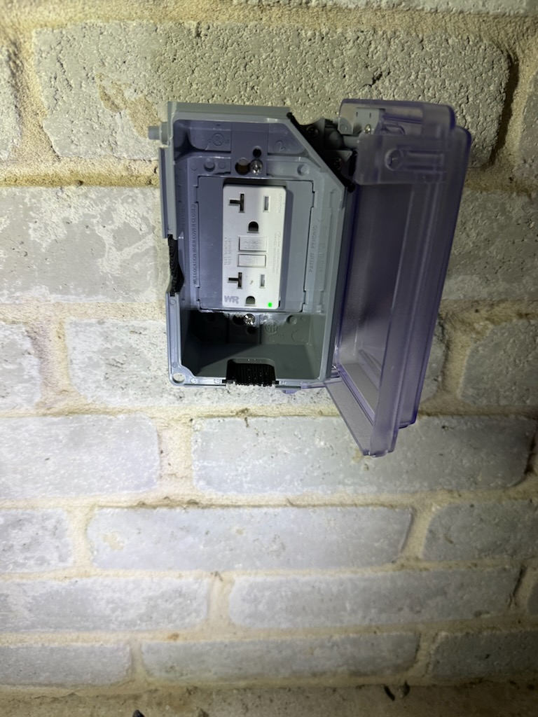 Updated outdoor outlet inside a gray weatherproof cover on a brick wall